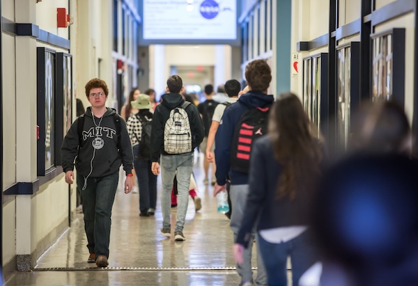 Busy hallway with blue walls and multiple people walking or talking; a person in an MIT hoodie walks toward the camera.